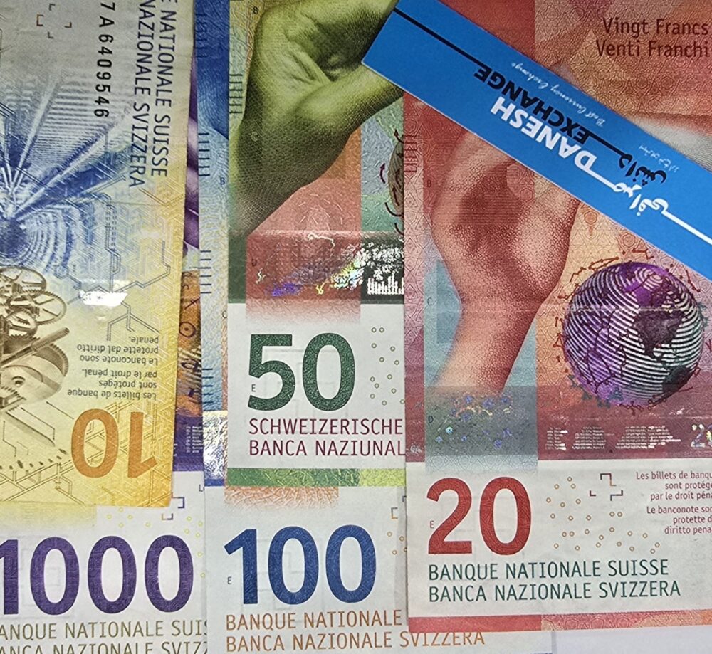 Assorted Swiss franc banknotes in multiple denominations, with a blue Danesh Exchange card partially visible on top. Assorted Swiss franc banknotes in multiple denominations, with a blue Danesh Exchange card partially visible on top.